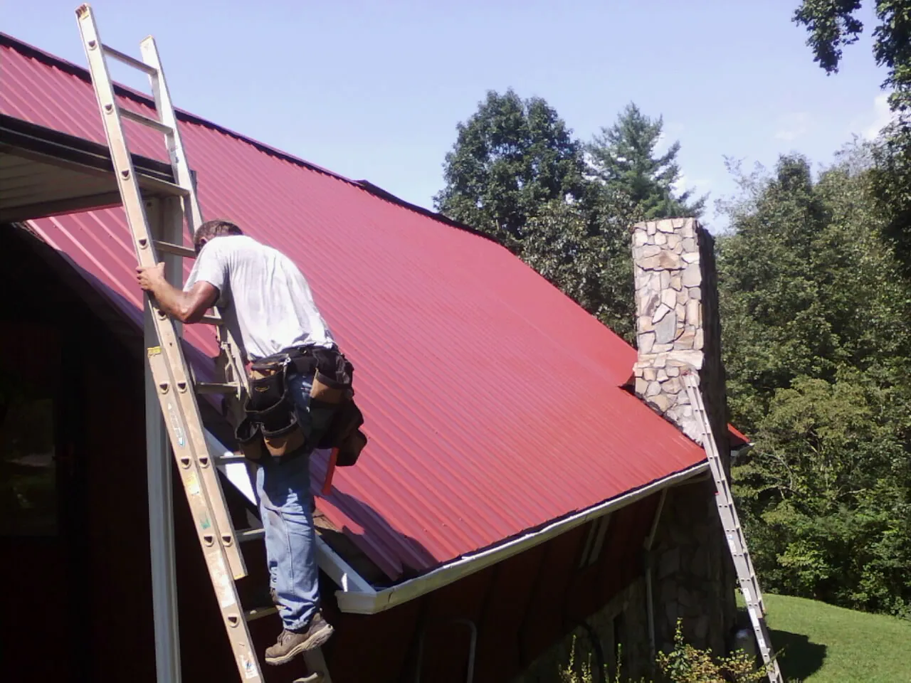 Roofer on a ladder beside a red metal roof and stone chimney — Soffit & Fascia Repair by Roofing Brothers in Garvin
